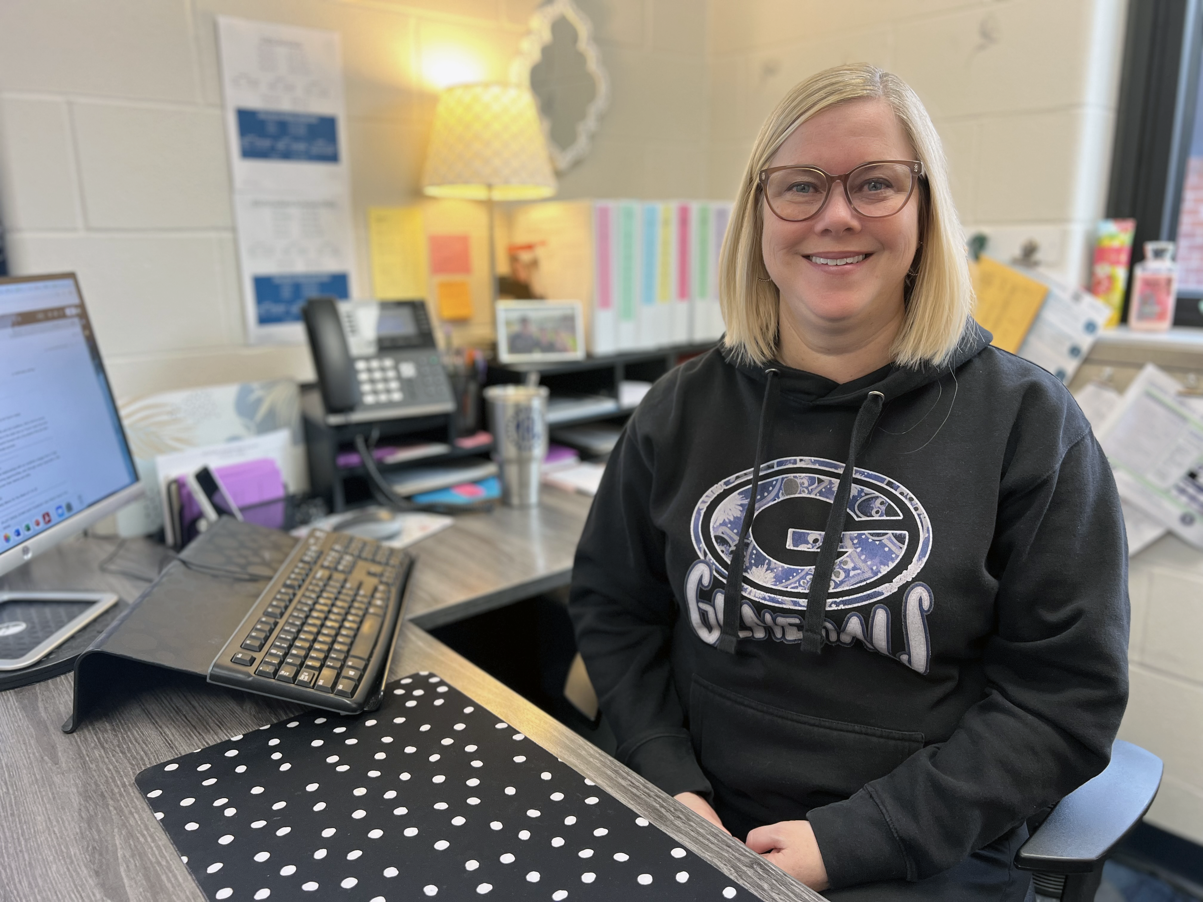 michelle reid is smiling in her office. She has blonde, shoulder-length hair and is wearing glasses and a black Generals sweatshirt