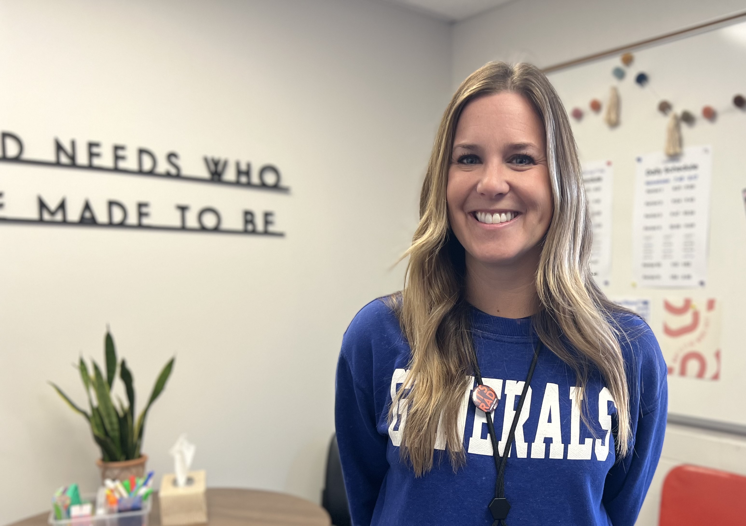 Shauna Dunbar, young woman with long blond hair smiles in her office. She is wearing a blue Generals sweatshirt. Behind her is a sign that reads, "the world needs who you were made to be"