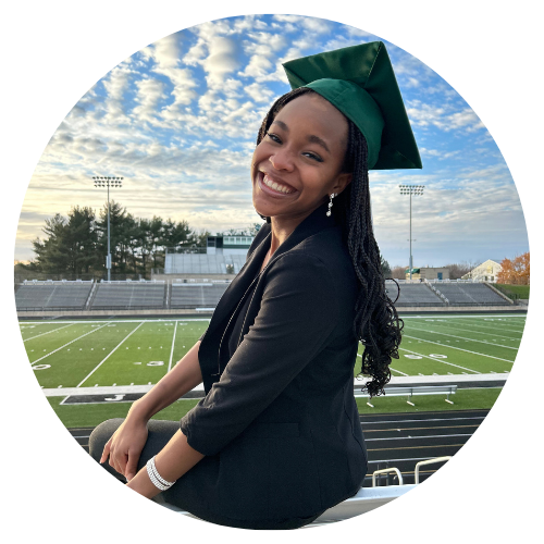 A medium-dark skin toned young women smiles toward the camera wearing a graduation cap, a black color shirt, grey pants, and a bracelet.  A football field and press box are located in the background.