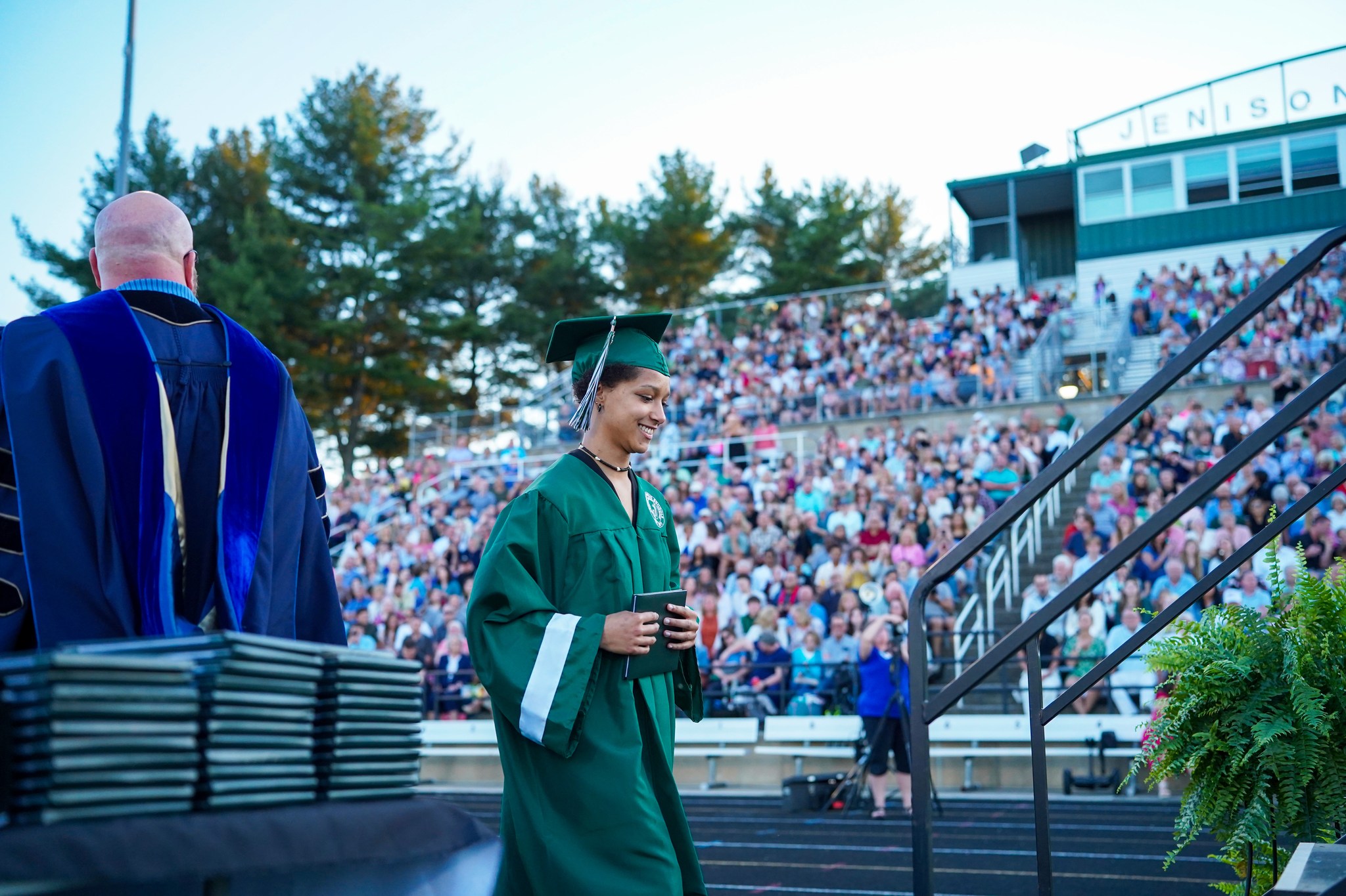 A JIA graduate receives her diploma, crossing the stage in cap and gown, with the crowd in the background.