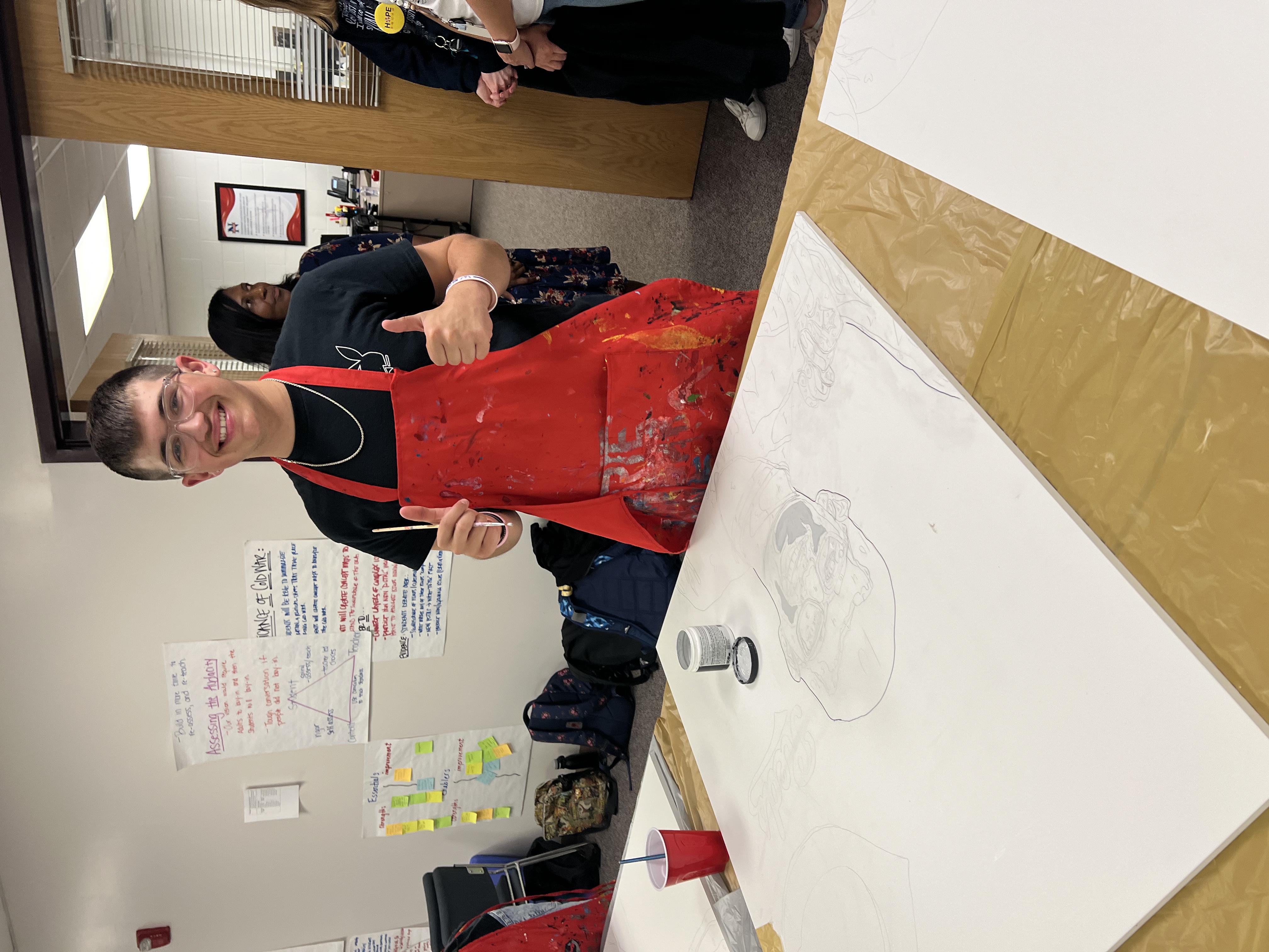 student wearing a painting apron. He is giving two thumbs up as he takes a break from painting his canvas. 
